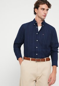 Tengerész kék gombos ing, fehér és piros függőleges csíkokkal, mellzsebbel és testre szabott fazonnal, világos bézs nadrággal és barna övvel párosítva.