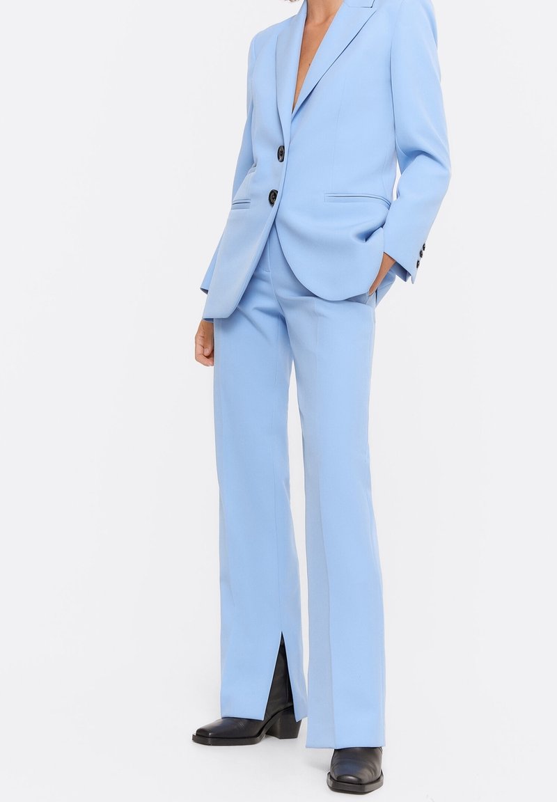 Woman wearing a light blue tailored suit with black boots, hands in pockets, standing against a plain white background.