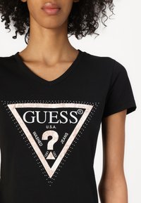 T-shirt nera a V con un motivo triangolare che presenta "GUESS®" e un punto interrogativo, decorato con strass e testo bianco.
