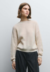 Maglione a maglia beige chiaro con scollo ampio, spalle scese e polsini a coste, abbinato a pantaloni scuri a righe. Texture morbida.