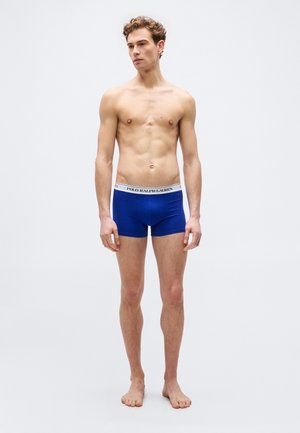 Mladić stoji bos, obučen u plave bokserice s bijelim pojasevima na kojima piše Polo Ralph Lauren, na jednostavnoj pozadini.