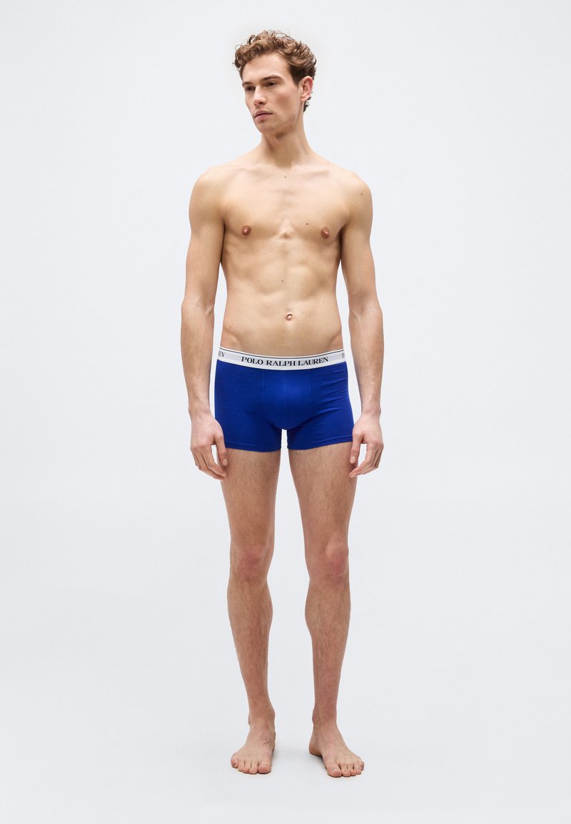 Νέος άνδρας στέκεται ξυπόλητος, φορώντας μπλε ανδρικά εσώρουχα τύπου boxer με λευκή ζώνη μέσης που φέρει την ετικέτα Polo Ralph Lauren, σε απλό φόντο.