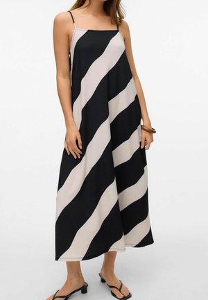 Femme portant une robe longue sans manches à rayures diagonales noires et blanches, avec bretelles fines et sandales noires à talons ouverts sur un fond uni.