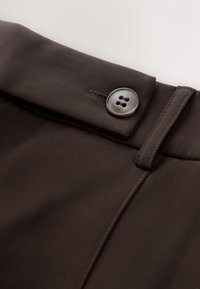 Pantalones de vestir marrones con una tela suave, líneas limpias y un botón decorativo de cuatro agujeros. Se puede ver una costura detallada.