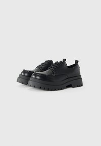 Par de zapatos de cuero negros con cordones, de suela gruesa tipo lug y detalles de alfiler metálico, sobre un fondo blanco.