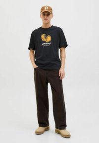 Schwarzes Baumwoll-T-Shirt mit orangefarbigem Grafikdruck, kombiniert mit lockeren dunklen Hosen und beigen Slippern. Die Mütze ist braun mit einem Logo.