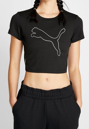 Femme portant un t-shirt court noir avec le logo Puma blanc et un pantalon noir assorti à taille élastique, les mains dans les poches.