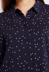 Blusa blu navy con piccoli motivi a cuori bianchi e beige, dotata di colletto e pattina con bottoni sullo scollo.