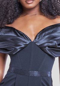 Donna che indossa un abito in satin nero con spalle scoperte, vita aderente e dettagli di tessuto arricciato sul petto.