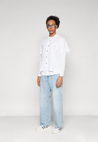 White short-sleeve shirt with eyelet pattern over a long-sleeve white top, paired with light blue wide-leg jeans and white sneakers.