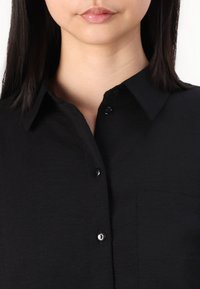 Black button-up shirt made of textured fabric, featuring a collar, front pocket, and black buttons on a single placket.