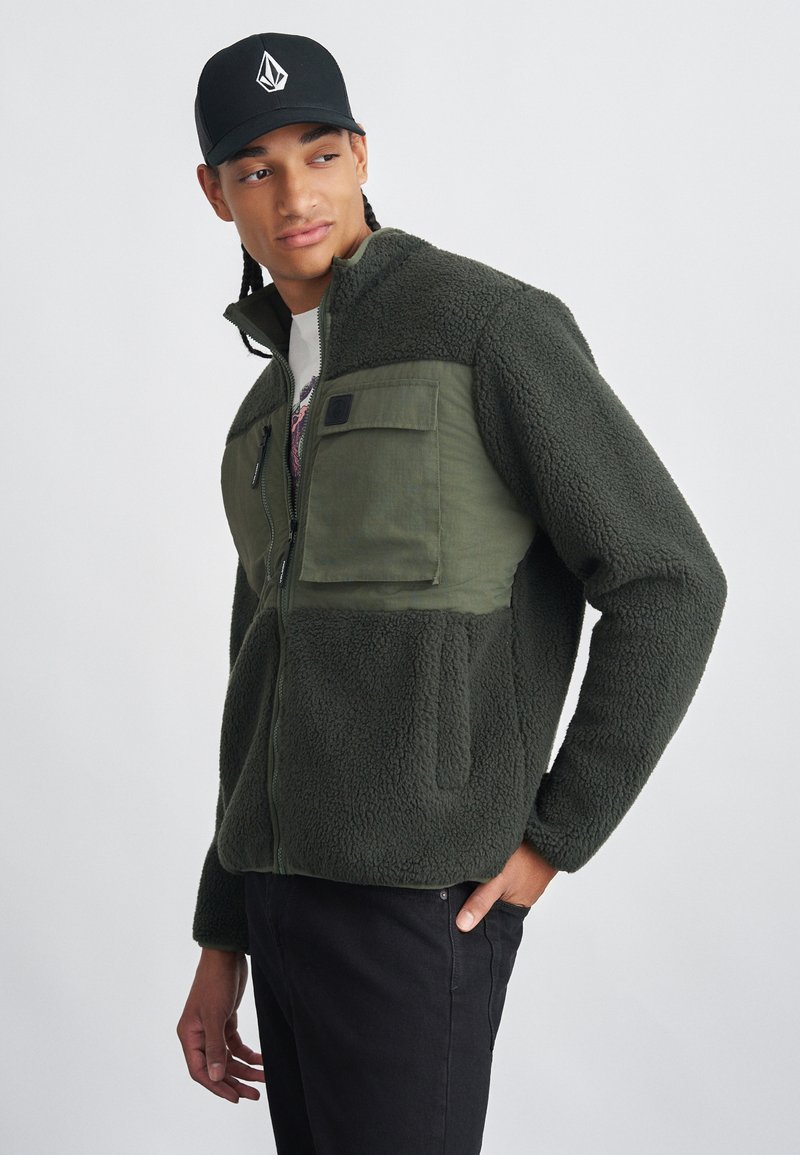 Fleece jacket in dark green with a textured, fluffy body and smooth fabric chest pockets; features a zip closure and high collar.