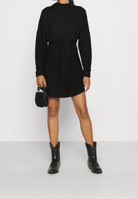 Robe noire à manches longues et taille ceinturée, avec un col haut. Portez avec des bottes de cowboy noires et un petit sac.