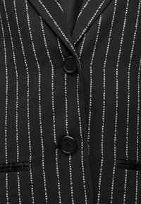 Μαύρο σακάκι με κάθετη λευκή εκτύπωση κειμένου που αναγράφει "KARL LAGERFELD", δύο κουμπιά μπροστά και μία τσέπη στο στήθος. Ομαλή υφή.