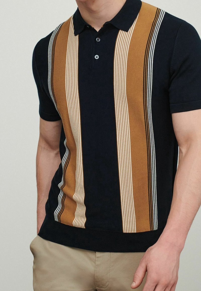 Homme portant un polo noir à manches courtes avec de larges rayures verticales marron, beige et blanches, associé à un pantalon beige.