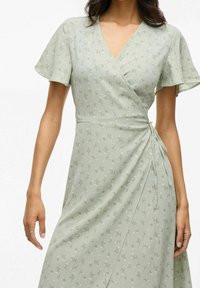 Kvinna i en ljusgrön omslagsklänning med korta volangärmar och små vita blommor, knuten i midjan på höger sida.