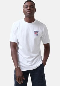 Camiseta de algodón blanca con un pequeño diseño impreso que dice "SURF", "ALOHA" y un gráfico colorido en la parte izquierda del pecho. Manga corta.