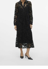 Abito camicia in pizzo nero con maniche lunghe, colletto e motivi floreali trasparenti. Lunghezza al di sotto del ginocchio, abbinato a scarpe nere con tacco basso.