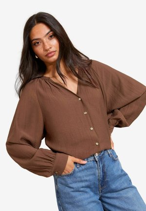 Braune, strukturierte Bluse mit V-Ausschnitt und Knopfverschluss. Weite Ärmel und dezente Raffungen an den Schultern. Kombiniert mit blauer Jeans.