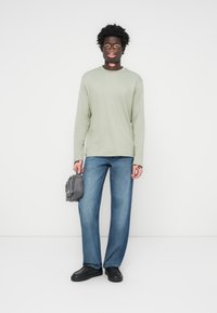 Lys grønne langermede skjorte, blå vide jeans, svarte sko, holder en grå veske. Enkel design med nøytrale farger og avslappet passform.