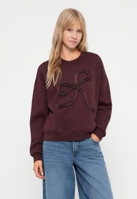 Sweatshirt bordeaux com um design de laço de fita texturizada na frente, apresentando um corte descontraído e punhos canelados, combinado com jeans azul claro.