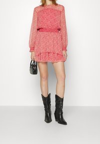 Robe rouge à motif floral avec manches longues en voile transparent, taille smockée et jupe à volants superposés. Associée à des bottes cowboy en cuir noir et un mini sac.