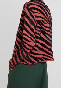 Maglione fuzzy in arancio ruggine con strisce di tigre nere, caratterizzato da un colletto rotondo e una vestibilità morbida, abbinato a pantaloni verde scuro.