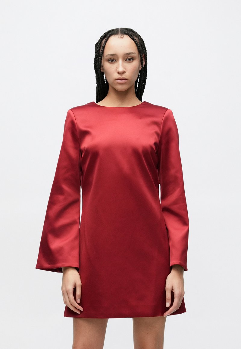 Kvinne med flettet hår iført en rød minikjole i sateng med lange ermer, mot en enkel hvit bakgrunn, vendt fremover med et nøytralt uttrykk.