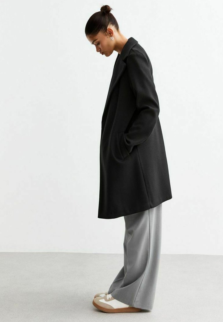 Manteau long noir au design ajusté, porté sur un pantalon large gris clair et des chaussures beiges avec des accents blancs. Texture lisse, poches latérales.