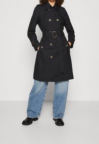 Svart trenchcoat i slät tyg, med bälte i midjan, dubbelknäppt design med stora knappar och breda slag.