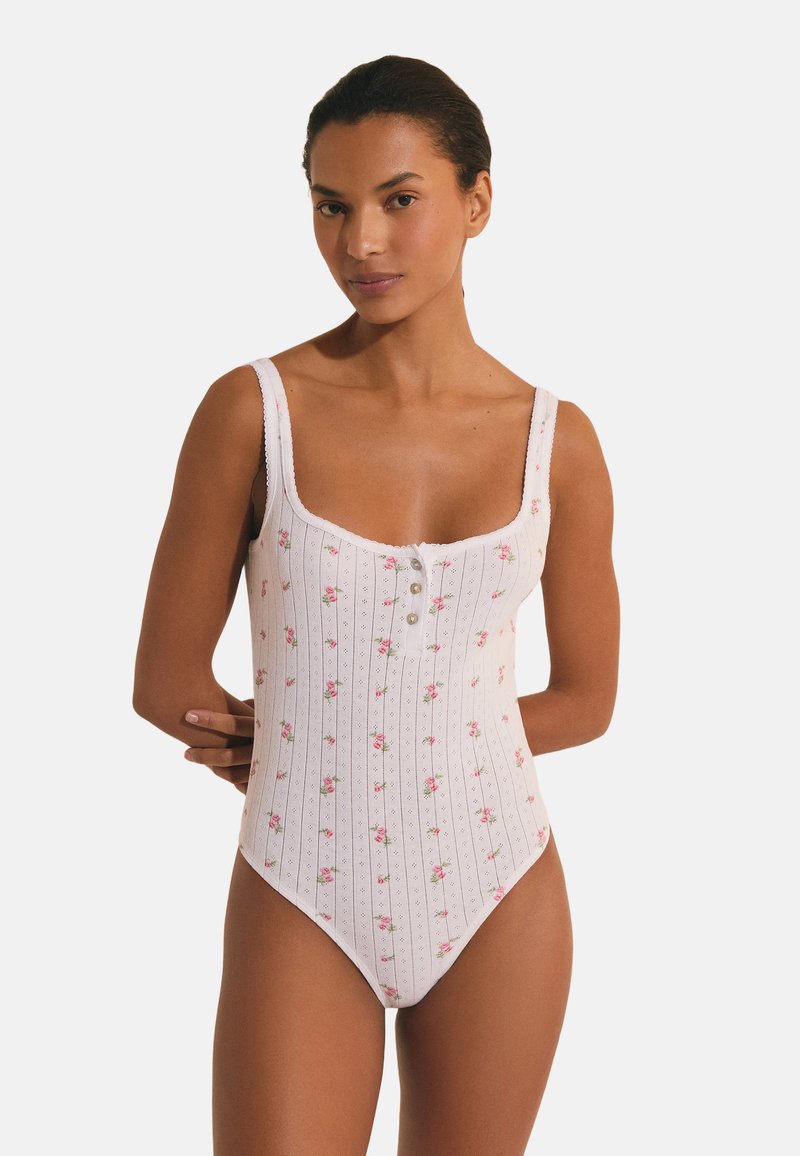 Frau trägt einen weißen, ärmellosen Bodysuit mit Blumenmuster und Knöpfen vorne, steht vor neutralem Hintergrund.