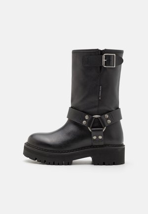 Bota negra de cuero hasta la mitad de la pantorrilla con detalles de hebilla y correa plateadas, suela robusta y cremallera lateral, mostrando una etiqueta con el logo de Tommy Jeans.
