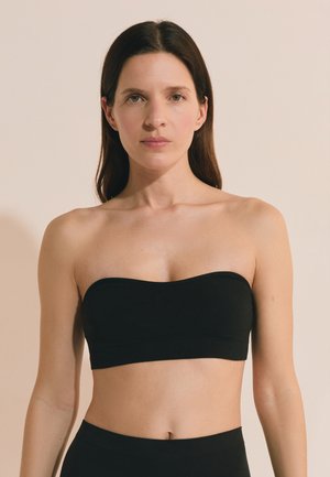 Woman with straight brown hair wearing a black strapless top, facing forward against a plain beige background.