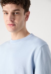 Světle modrý svetr vyrobený z měkké tkaniny, s kulatým výstřihem a žebrovanými manžetami, s malým bílým logem "BOSS" na hrudi.