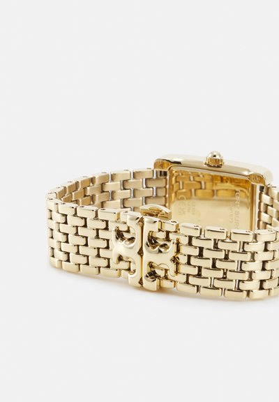 Goudkleurige metalen horlogeband met een rechthoekige kast en een decoratieve logospeld, getoond van achteren en van de zijkant op een witte achtergrond.