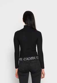 Svart ribbad turtlenecktröja med långa ärmar, figurnära design och en vit märkestappning. Bärs med svarta högmidjade jeans.