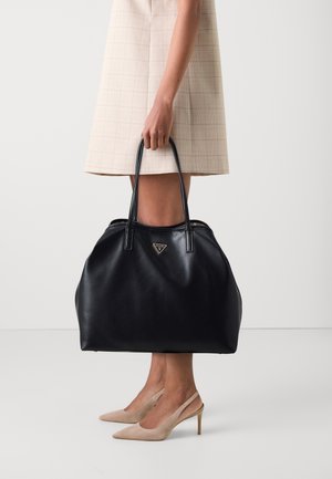 Sac fourre-tout en cuir noir de forme arrondie, présentant un logo triangulaire en or, associé à une jupe plissée claire à carreaux et des chaussures à talons hauts couleur nude.