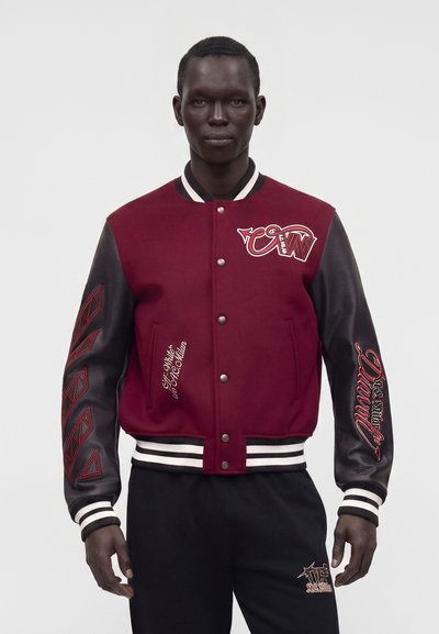 Homme portant une veste universitaire bordeaux et noire avec du texte brodé sur les manches et la poitrine, debout devant un fond clair uni.