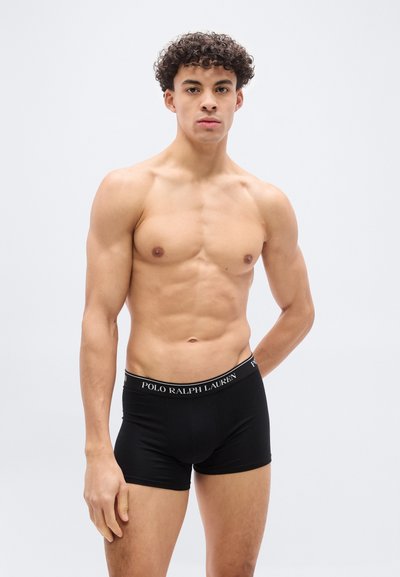 Μυώδης νεαρός με σγουρά μαλλιά, φορώντας μαύρα εσώρουχα μπόξερ Polo Ralph Lauren, στέκεται μπροστά σε απλό λευκό φόντο.