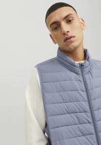 Young man with short hair wearing a light blue quilted vest over a white long-sleeve shirt, looking slightly downward against plain background.