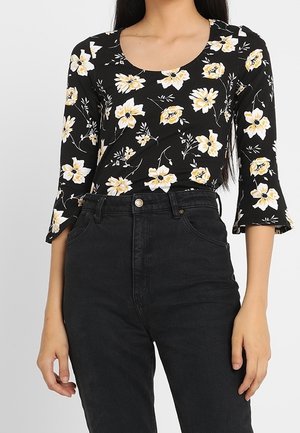 Top noir ajusté avec imprimé floral jaune et blanc, manches trois-quarts, associé à un jean noir taille haute.
