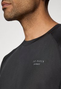Černé sportovní tričko s kulatým výstřihem a raglánovými rukávy. Na hrudi je bílé logo "Ted Baker Sport". Hladká textura.