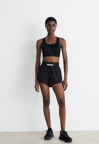 Persona snella che indossa un reggiseno sportivo nero, pantaloni corti neri Calvin Klein e sneakers nere, in piedi contro un muro bianco semplice.