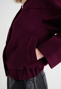 Veste bordeaux avec fermeture éclair, taille côtelée, poches latérales et tissu texturé. Le design présente des manches larges.