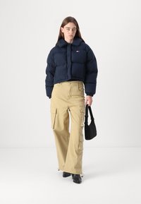 Marineblauwe cropped pufferjack met hoge kraag, beige cargo pants en zwarte haklaarzen. Klein logo detail op de jas. Zwarte tas.