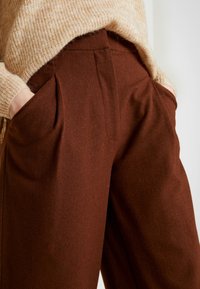 Handen in de zakken van een hoog-wijd, bruine broek, gecombineerd met een beige gebreide trui tegen een neutrale achtergrond.