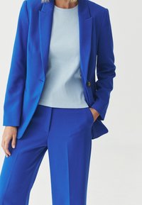 Modrý látkový oblek sestávající z ušitého saka s jedním knoflíkem a sladěných kalhot, nošený přes světle modrý top.