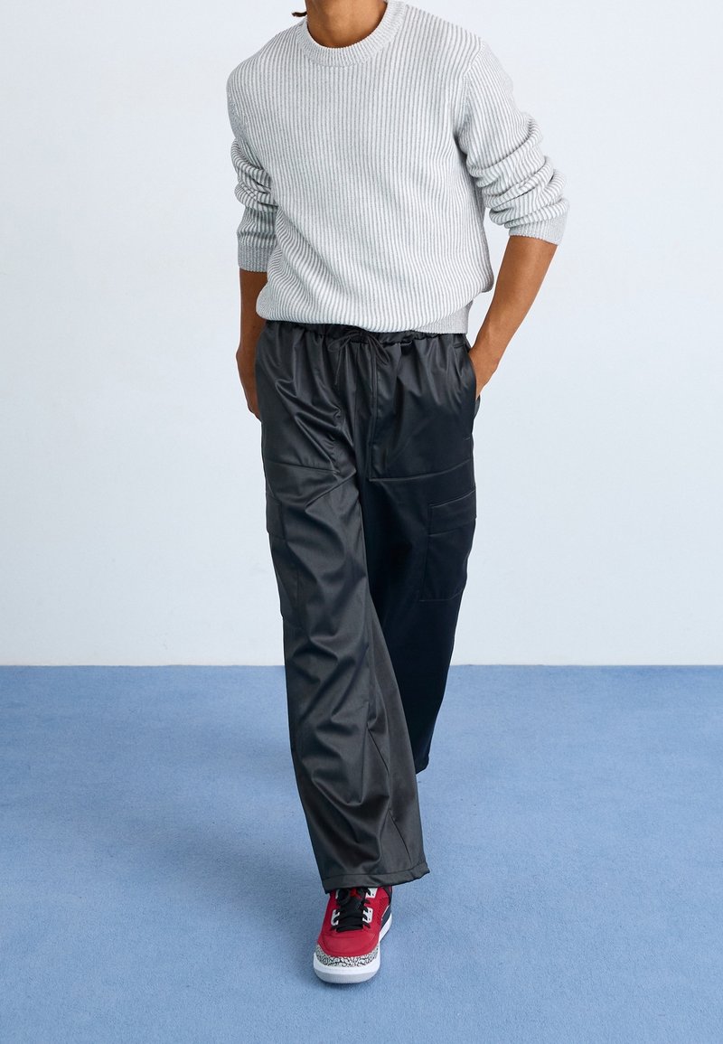 Personne portant un pull en maille gris clair, un pantalon cargo noir avec cordon de serrage et des baskets rouges, debout sur un sol bleu contre un mur blanc.