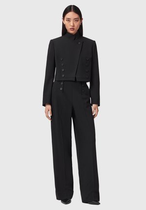 Femme portant un blazer court à double boutonnage noir assorti à un pantalon taille haute à jambes larges et des chaussures noires pointues.