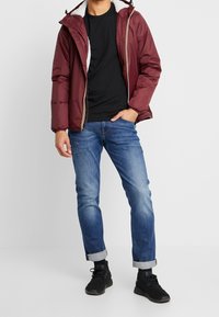 Persona che indossa una giacca bordeaux con fodera in pelliccia bianca, t-shirt nera, jeans blu arrotolati e sneaker nere, in piedi contro uno sfondo semplice.